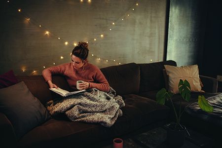 A woman sits on a sofa, reading a book under a blanket in a comfy looking room.