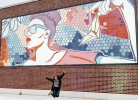 Marissa jumps into the air in front of her Reotown mural, which depicts a stylish woman looking at a paper crane dangling from her finger