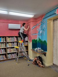 Marissa sits on top of a bookshelf while she paints on the wall
