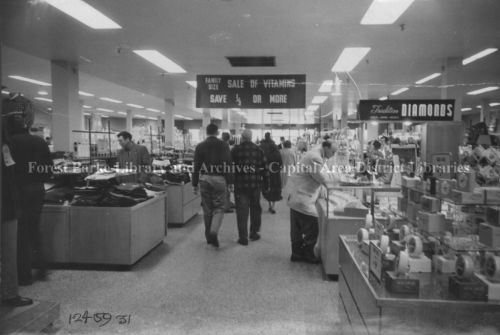 Black and white photograph of interior of department store. Shoppers browsing displays.