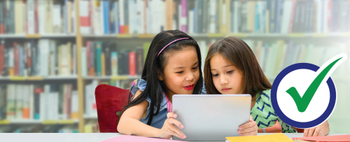 Two young girls reading from a tablet with bookshelves in the background. Checkmark icon.